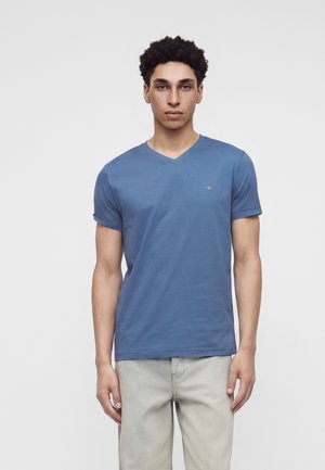 Jeune homme aux cheveux bouclés portant un T-shirt bleu col en V et un pantalon gris clair, debout sur un fond blanc uni.