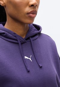 Μωβ φούτερ με κουκούλα, φτιαγμένο από μαλακό ύφασμα, με λευκό λογότυπο Puma, κουκούλα με κορδόνι και ριμπ λεπτομέρειες κοντά στον λαιμό και τις μανσέτες.