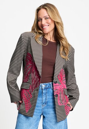 Lachende vrouw met lang blond haar draagt een geruite blazer met rode bloemenborduursel over een bruin topje en een hooggesloten blauwe spijkerbroek.