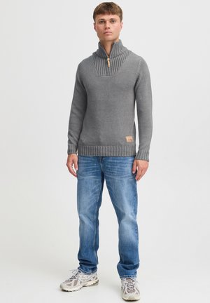Grå strikket sweater med høj krave, ribbede detaljer og et lille logo-patch ved kanten, parret med blå jeans og træningssko.