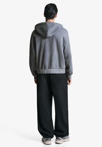 Muž stojaci chrbtom, oblečený v šedej mikine, voľných čiernych nohaviciach a šedých teniskách na bielom pozadí.
