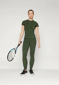 Olive rohelise värvi sportlik särk ja legginsid, mis on valmistatud venivast kangast, koos mustade spordijalatsitega. Isik hoiab tennise reketti.