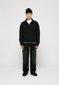 Schwarzer Strickpullover mit Kragen, über einem weißen Hemd getragen, kombiniert mit schwarzen Hosen mit Lederakzenten und silbernen Beschlägen.