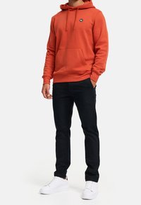 Orange hoodie med dragsnören och framficka, i kombination med svarta byxor och vita sneakers. Hoodien har en liten logotyp på bröstet.