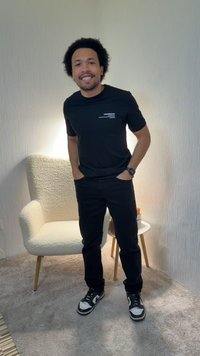 Afros frizurájú férfi mosolyog, fekete pólót és nadrágot visel, zsebében tartott kézzel áll egy fehér, texturált karosszék mellett, mellett egy kis kerek asztal.