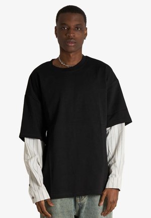 Giovane uomo che indossa una T-shirt nera oversize semplice sopra una camicia bianca a maniche lunghe con righe verticali, in piedi davanti a uno sfondo bianco.