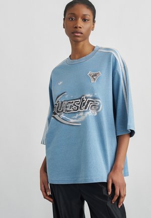 Ung kvinde iført oversize blå sportst-shirt med hvide striber på ærmerne og grafisk tekst på brystet, stående mod en ensfarvet baggrund.