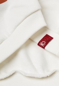 Felpa bianca con polsini a costine, dotata di una morbida fodera interna. Dettaglio significativo: un'etichetta rossa glitterata con un accento di logo bianco.