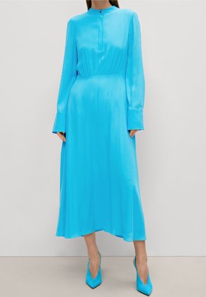 Kvinde iført en langærmet, lyseblå midi-kjole med høj krave og matchende spidse hæle mod en ensfarvet baggrund.