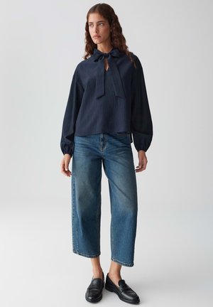 Navyblaue Bluse mit gebundenem Ausschnitt und strukturiertem Stoff, kombiniert mit lässigen, hellblauen, cropped Jeans und schwarzen Slippers.