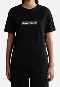Persona que lleva una camiseta negra de manga corta con el logo blanco "NAPAPIJRI geographic" en el pecho, combinada con pantalones negros.