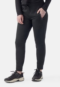 Pantalones jogger negros ajustados en un tejido suave y elástico, con cintura con cordón y bolsillos laterales con cremallera. Combinados con zapatillas deportivas negras.