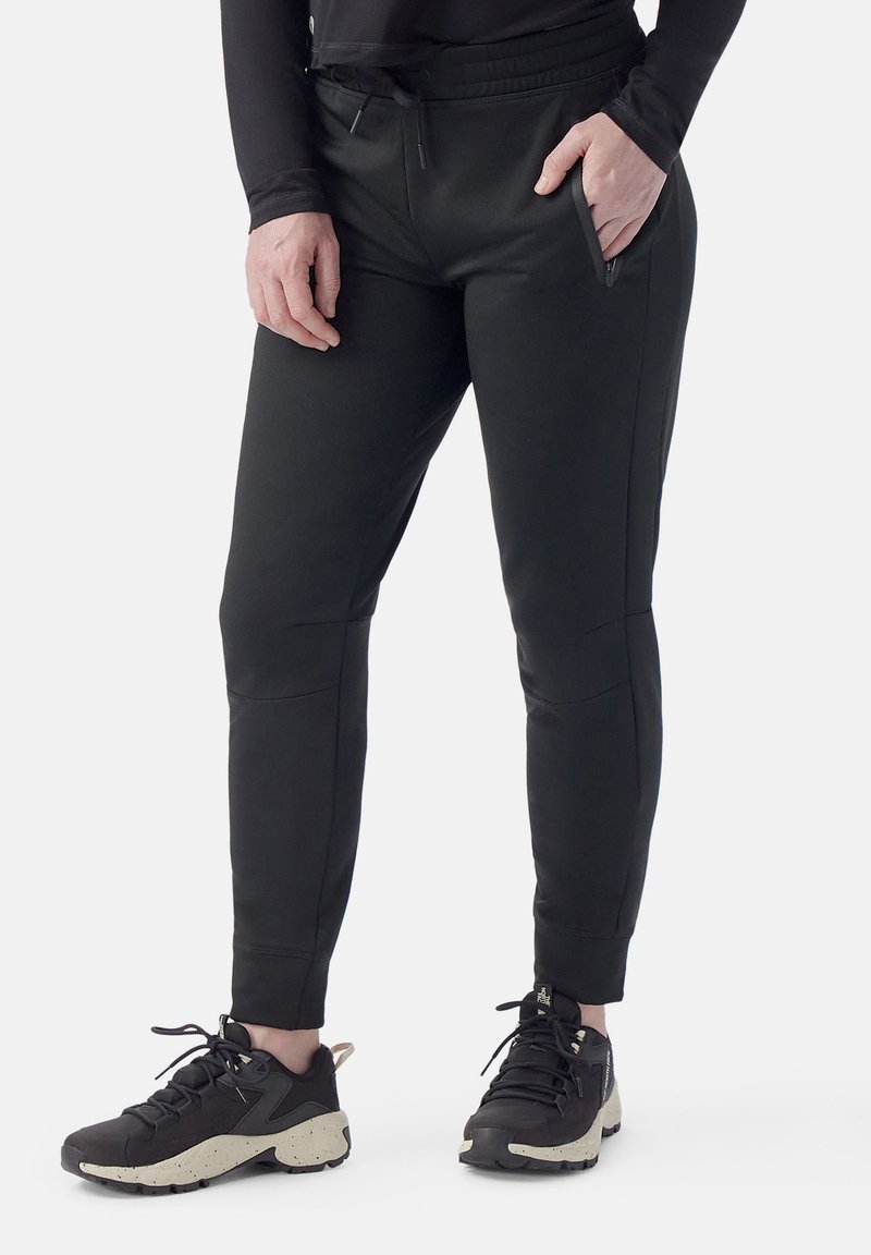 Pantalones jogger negros ajustados en un tejido suave y elástico, con cintura con cordón y bolsillos laterales con cremallera. Combinados con zapatillas deportivas negras.