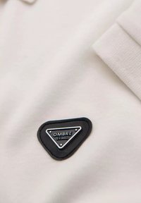 Hvid polo shirt stof med en tekstureret overflade, der har en sort trekantet læderlap prægede med "OMBRO EXIMIO."