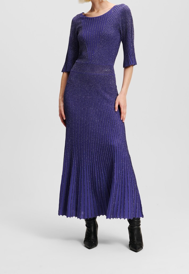 Femme portant une robe midi plissée violette scintillante avec des manches mi-longues jusqu'au coude et des bottines noires à bouts pointus.