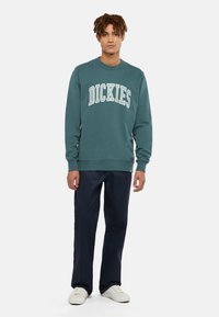 Teal sweatshirt med "DICKIES" tryckt i vit bågformad text. Har en rund hals, långa ärmar och ribbad mudd. Bärs med marinblå byxor.
