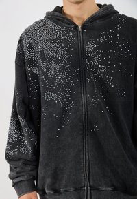 Sweatshirt negru cu glugă, prevăzut cu fermoar, decorat cu un model dispersat de piepteni albe pe partea din față superioară. Textură moale.