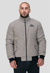 Veste matelassée à motifs à carreaux dans des tons beige et marron avec un col haut, une fermeture éclair, et des accents noirs au niveau des poignets et de l'ourlet.