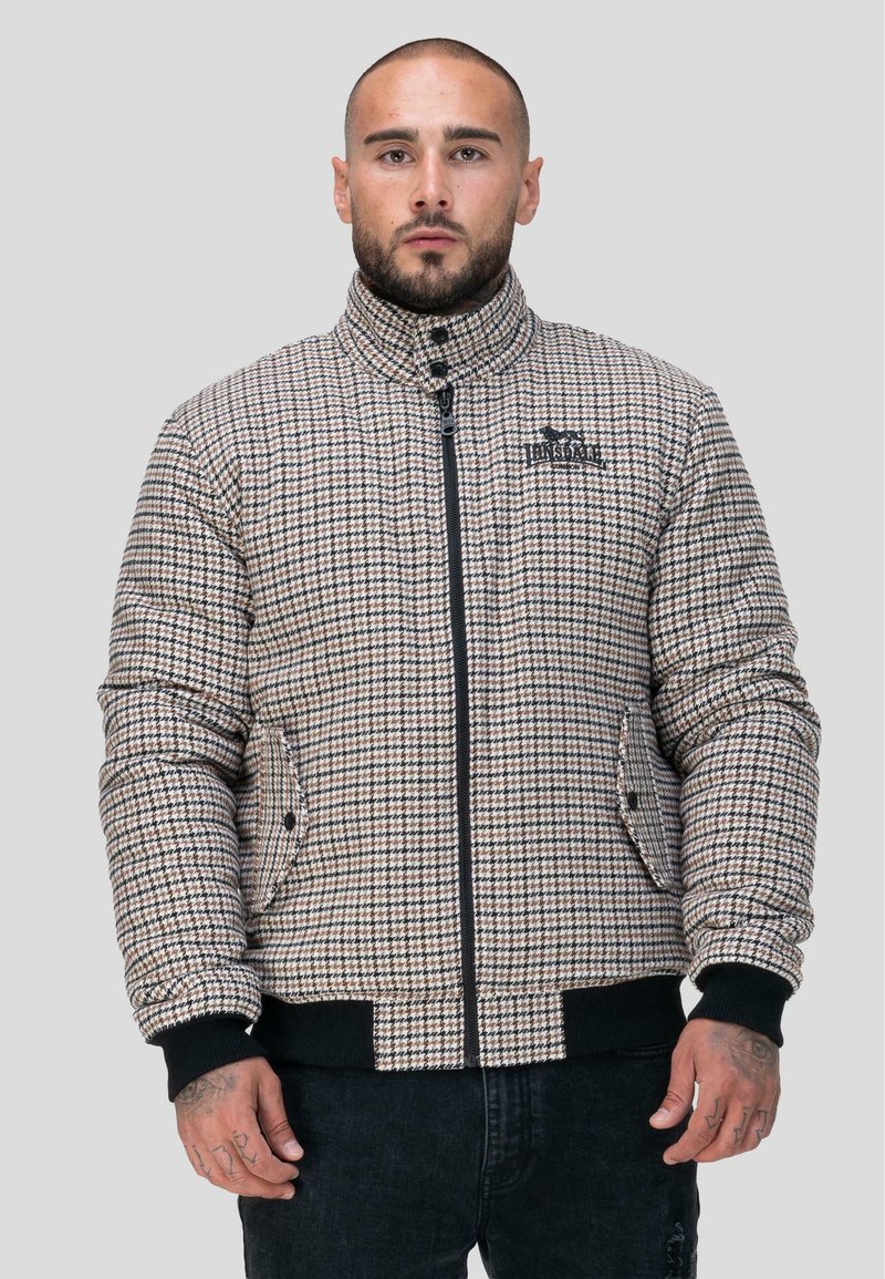 Veste matelassée à motifs à carreaux dans des tons beige et marron avec un col haut, une fermeture éclair, et des accents noirs au niveau des poignets et de l'ourlet.