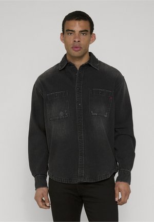 Jonge man draagt een vervaagd zwart denim overhemd met twee borstzakken, staand tegen een effen grijze achtergrond.