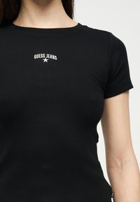 Μαύρο κοντομάνικο μπλουζάκι με ραβδώσεις, φορεμένο από άτομο, με λευκό κείμενο «GUESS JEANS» και λογότυπο αστεριού στο κέντρο του στήθους.