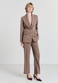 Brauner, maßgeschneiderter Anzug mit einreihigem Blazer, der goldene Knöpfe, zwei Taschen und eine schmal geschnittene Hose umfasst. Elegante burgunderrote High Heels vervollständigen das Outfit.