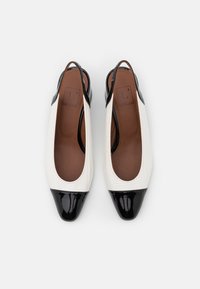 Bílé klínové boty s texturovaným povrchem, černé lakované kožené akcenty na špičce a hnědá vnitřní podšívka.