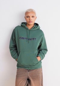 Hanorac verde din bumbac, având o buzunar frontal și glugă cu șnur, cu logo-ul purpuriu "Carhartt" pe piept.