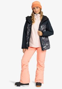 Navy-blaues Schneejacke mit farbenfrohem Grafikdesign, kombiniert mit einem hellrosa Fleece und korallenfarbigen Skihosen. Das Modell trägt eine gerippte orange Mütze.