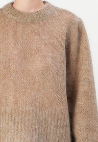 Primo piano di una persona che indossa un morbido e peloso maglione lavorato a maglia di un colore marrone chiaro, con scollatura rotonda a coste e polsini a coste.