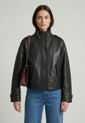 Frau trägt schwarze Lederjacke, blaue Jeans und burgunderfarbene Schultertasche mit Kettenriemen, steht vor einem schlichten hellen Hintergrund.