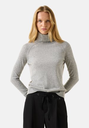 En kvinde med blondt hår bærer en lysegrå rullekravesweater og sorte bukser med snøre, og står med hænderne bag ryggen.