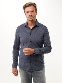Uomo che indossa una camicia a righe verticali blu navy e bianche con bottoni e jeans grigio chiaro, in piedi contro uno sfondo bianco semplice.