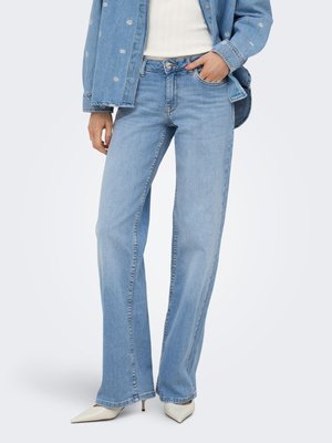 Personne portant un jean bleu clair à jambes droites, des talons blancs pointus, un haut blanc et une veste en jean bleue avec de petits motifs floraux.