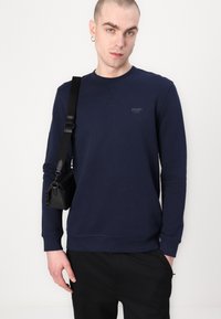 Sudadera de algodón azul marino con mangas largas, cuello redondo y un sutil logo "JOOP! JEANS" en el pecho. Bolsa de tirante negra en el hombro.