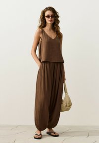 Femme aux cheveux ondulés portant un haut marron sans manches et un pantalon ample, lunettes de soleil foncées, sandales noires, tenant un sac à main beige, debout sur un sol carrelé.