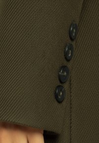 Olijfgroene stof met geribbelde textuur, met vier zwarte knopen langs de rand. Subtiele stikdetails versterken de structuur van het kledingstuk.