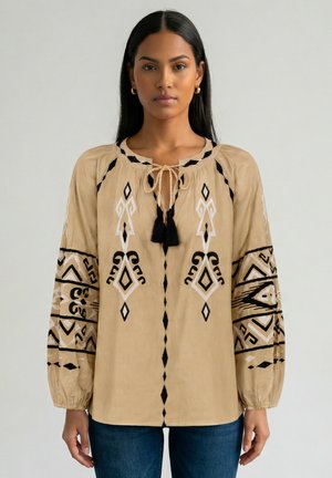 Vrouw met lang zwart haar draagt een beige blouse met zwart-witte geometrische borduring en tassels, gecombineerd met blauwe spijkerbroek.
