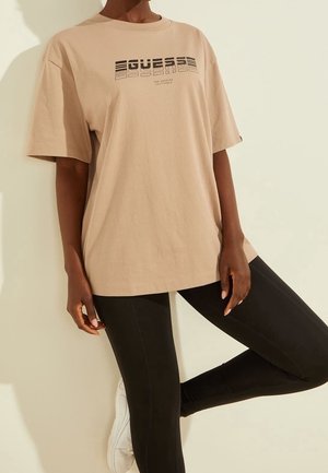 Personne portant un t-shirt beige ample avec le texte "GUESS Los Angeles California", des leggings noirs et des baskets blanches, en position détendue.