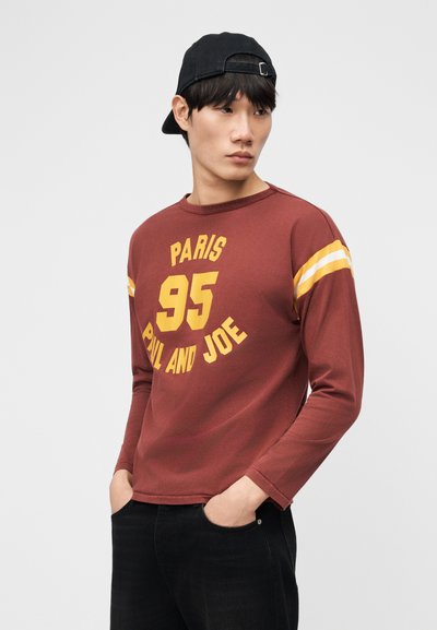 Jeune homme portant une casquette noire à l'envers et une chemise à manches longues bordeaux avec le texte « Paris 95 Paul and Joe » et des rayures jaunes sur les manches.