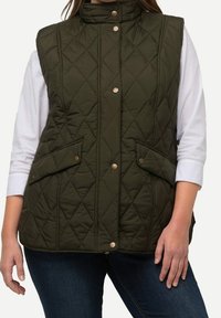 Olivgrüne gesteppte Weste mit Druckknöpfen und Klappentaschen, getragen über einem weißen Langarmshirt und dunkelblauen Jeans.