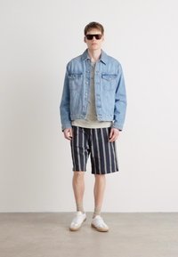 Veste en denim bleu clair, associée à un t-shirt gris et un short à rayures noir et blanc. Baskets blanches avec semelles beiges. Lunettes de soleil portées.