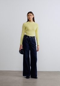 Κίτρινο πουλόβερ με ανάγλυφη πλέξη, υψηλός λαιμός, συνδυασμένο με σκούρο μπλε τζιν με φαρδιά μπατζάκια, μαύρη ζώνη, κρατώντας μαύρο φάκελο, με ομαλή υφή.