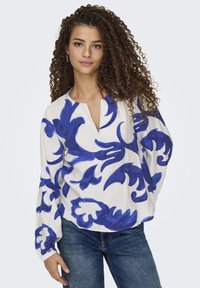 Weißbluse mit blauen abstrakten Blumenmustern, V-Ausschnitt, langen Ärmeln, fließendem Stoff, kombiniert mit blauen Jeans.