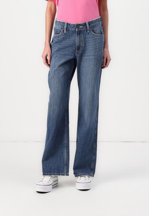 Person wearing mid-rise, straight-leg blue jeans with frayed hems, white platform sneakers, and a pink short-sleeve top standing against white background.