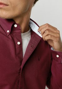 Camisa de botones burdeos con un cuello a rayas en azul y blanco que contrasta. Presenta botones blancos y un logo bordado en el pecho.