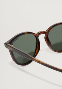 Sunglasses od kornjačevine s zelenim lećama, zaobljenog oblika i elegantnim crnim akcentima. Zlatni logo "Polaroid" na okviru.