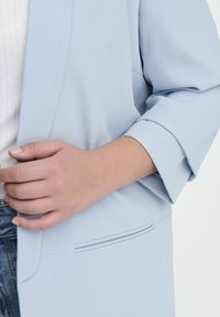 Hele sinine bleiser pehmest kangast, millel on kohandatud disain, kergelt rullitud varrukad ja ühel küljel õmmeldud detailiga tasku.