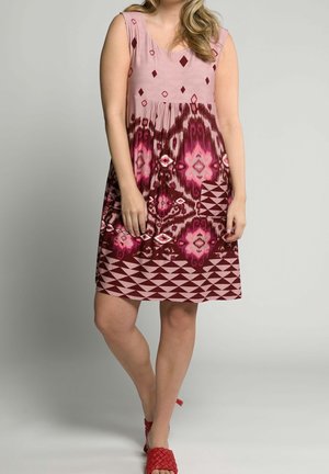 Frau in ärmellosem, knielangem Kleid mit pink- und kastanienfarbenem Muster und roten, flachen gewebten Schuhen, die vor einem schlichten Hintergrund steht.