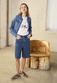 Femme debout les mains dans les poches, portant une veste en jean, un t-shirt blanc à motif, un short en jean et des baskets marron, à côté d'une chaise en bois rustique.